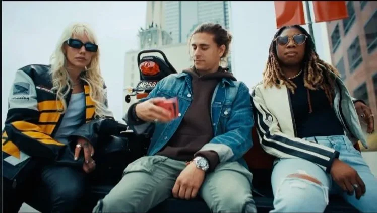 Three young adults sitting outdoors with a cityscape in the background, two women and one man, dressed casually, with one woman wearing sunglasses and a black and yellow jacket, the man looking at his phone, and the other woman wearing sunglasses and