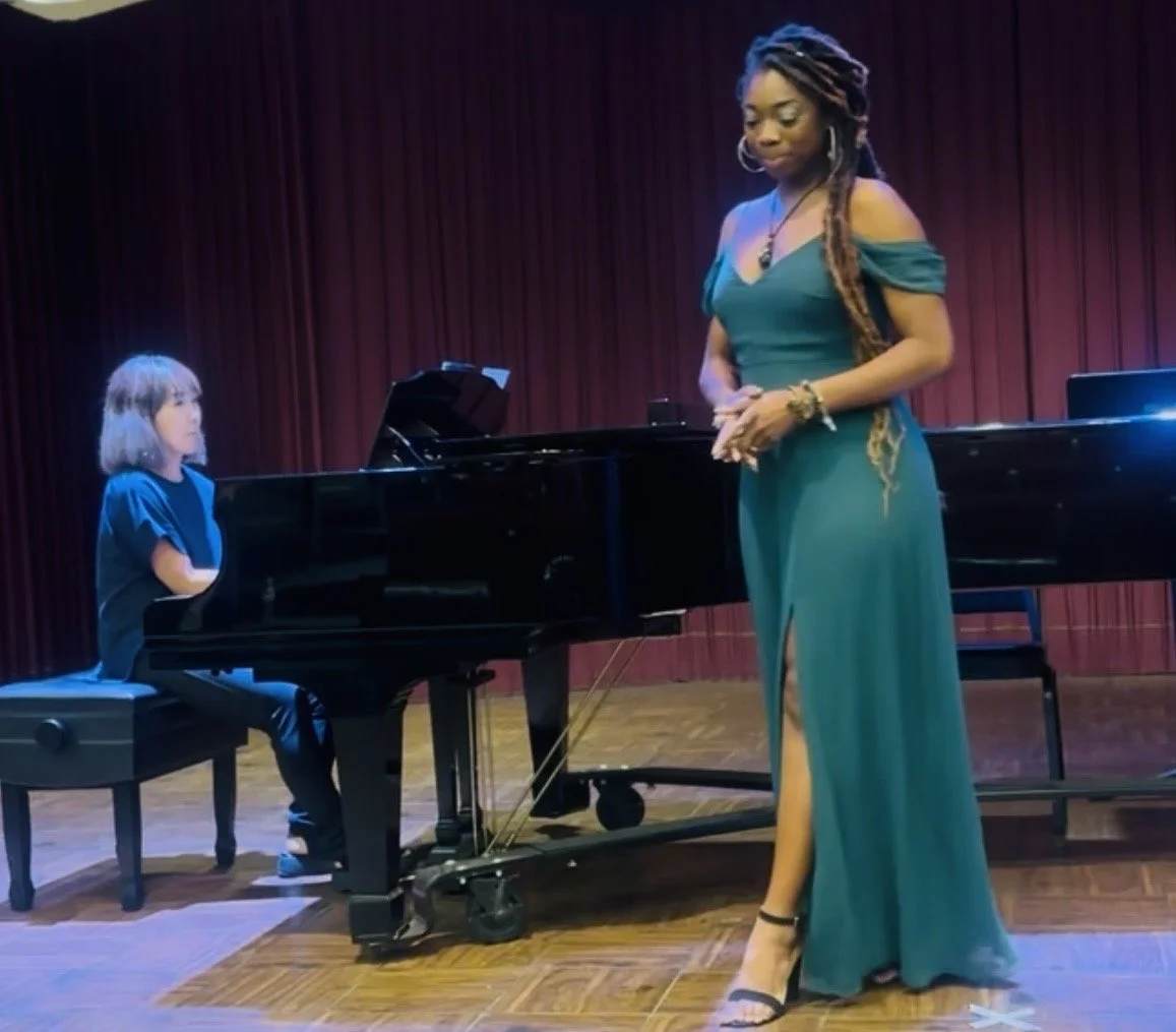 A woman in a green gown standing on stage, with a girl playing the piano next to her, red curtain backdrop.