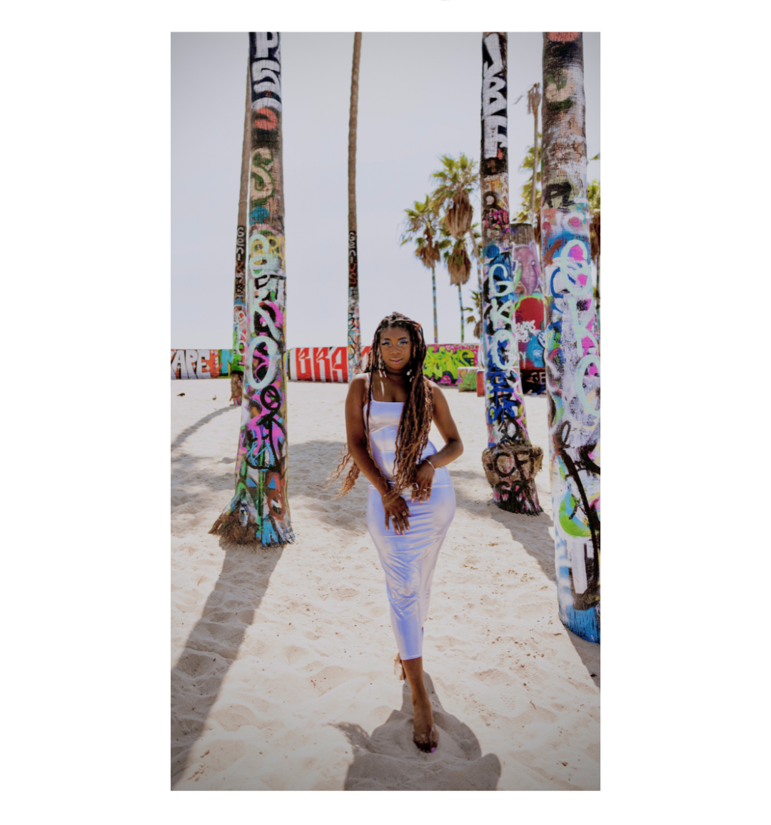 Woman in white dress standing on sandy beach between colorful graffiti-covered wooden posts, with palm trees and bright sky in background.