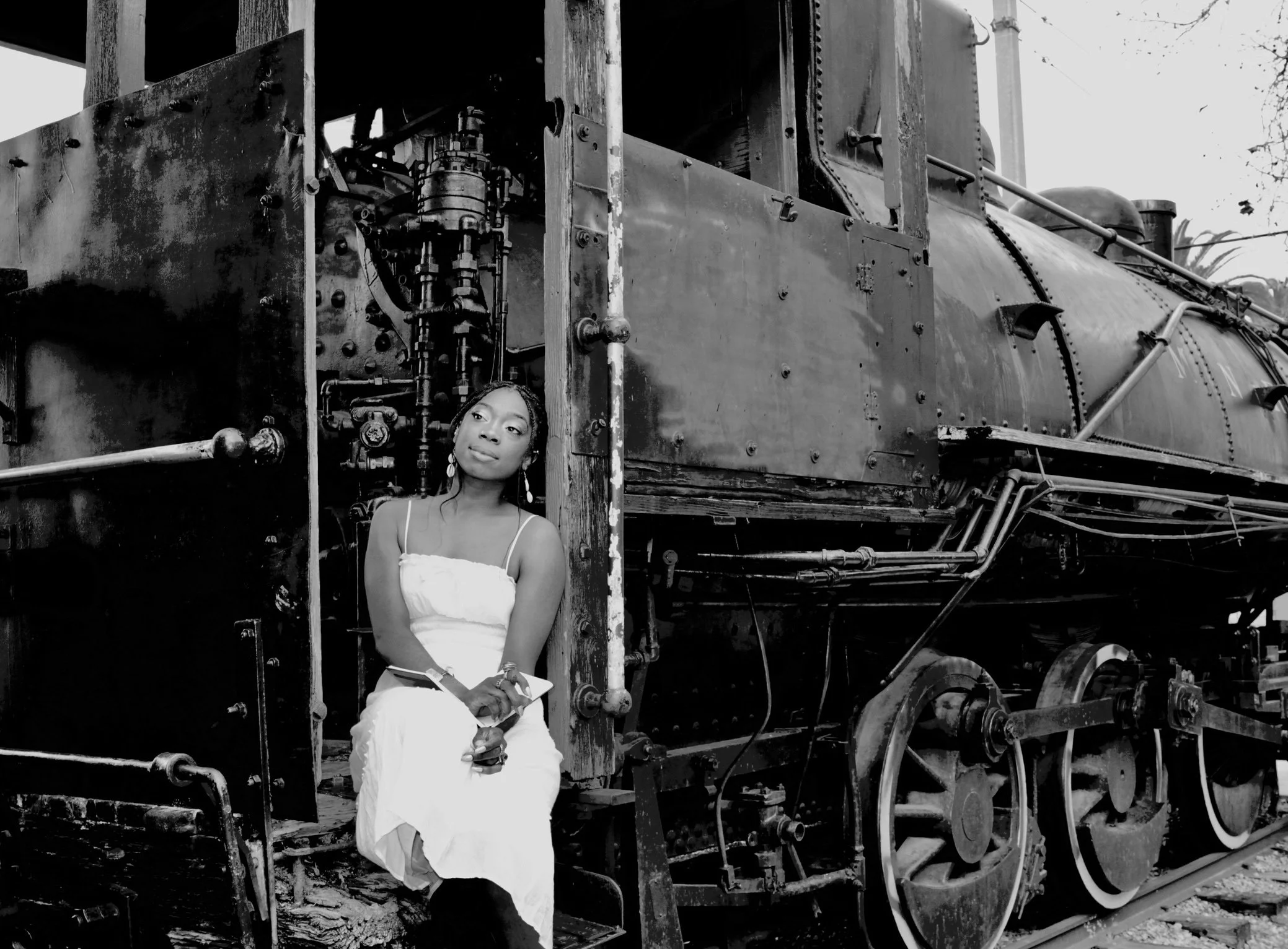 A woman in a white dress sits on the edge of an old steam locomotive, looking pensively to the side, with the large wheels and mechanical parts of the train nearby.