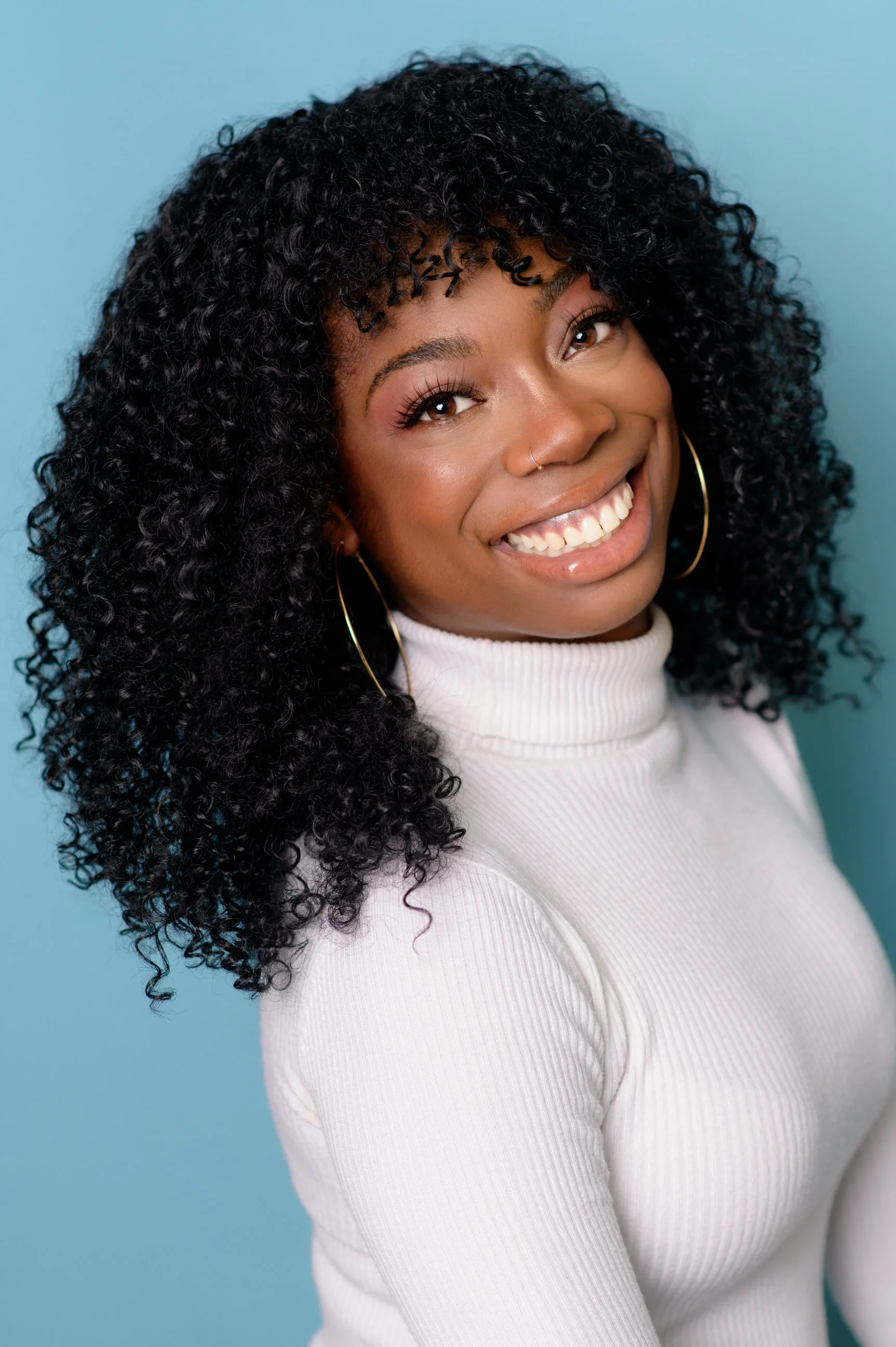 A young woman with curly black hair, smiling, wearing a white turtleneck sweater and large hoop earrings, against a light blue background.