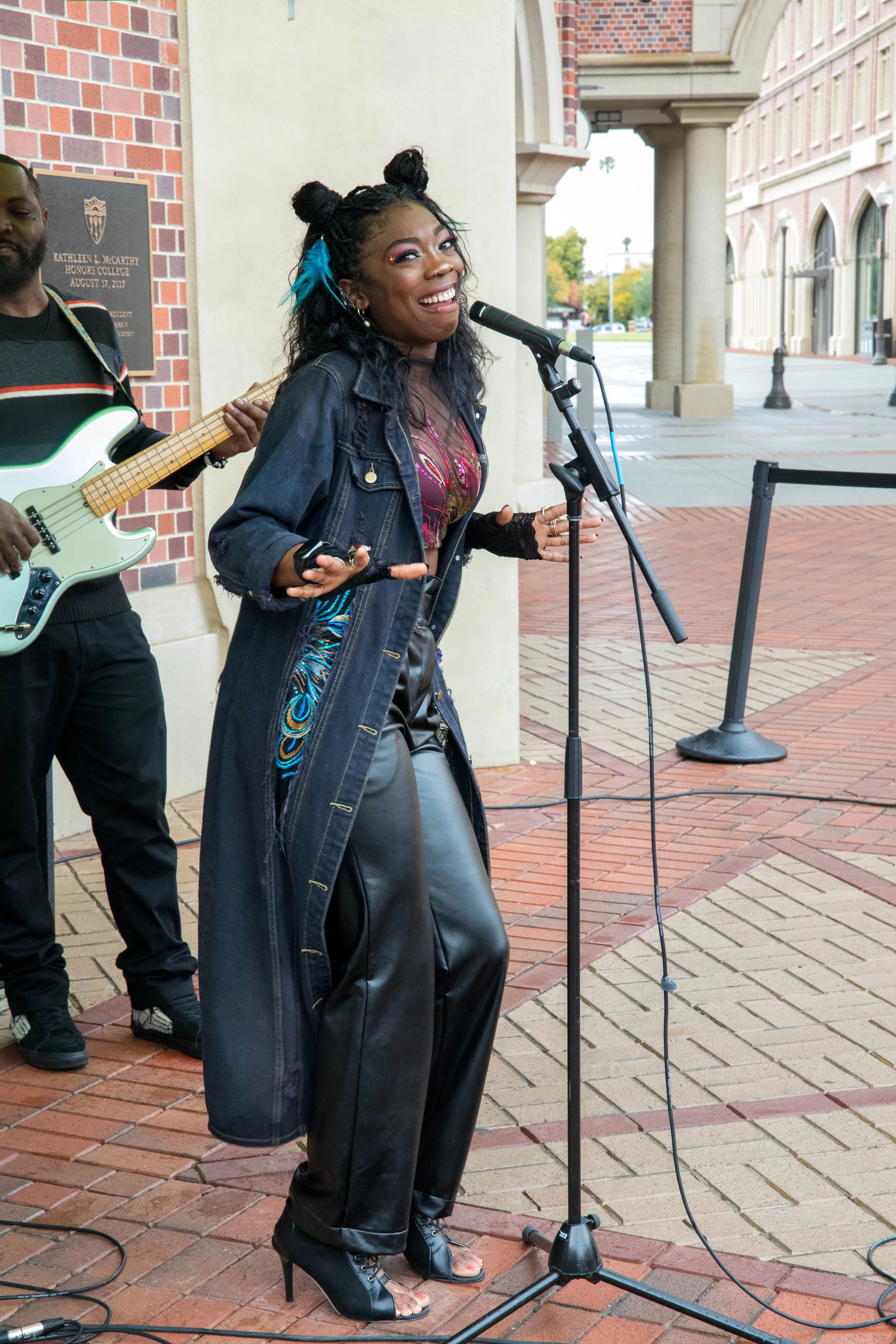 A woman with styled hair, wearing a dark denim coat, and black leather pants, singing into a microphone on a stand outdoors with a man playing guitar beside her.