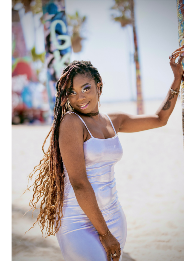 A woman with long dreadlocks wearing a satin white dress at the beach, smiling and posing near a colorful pole.