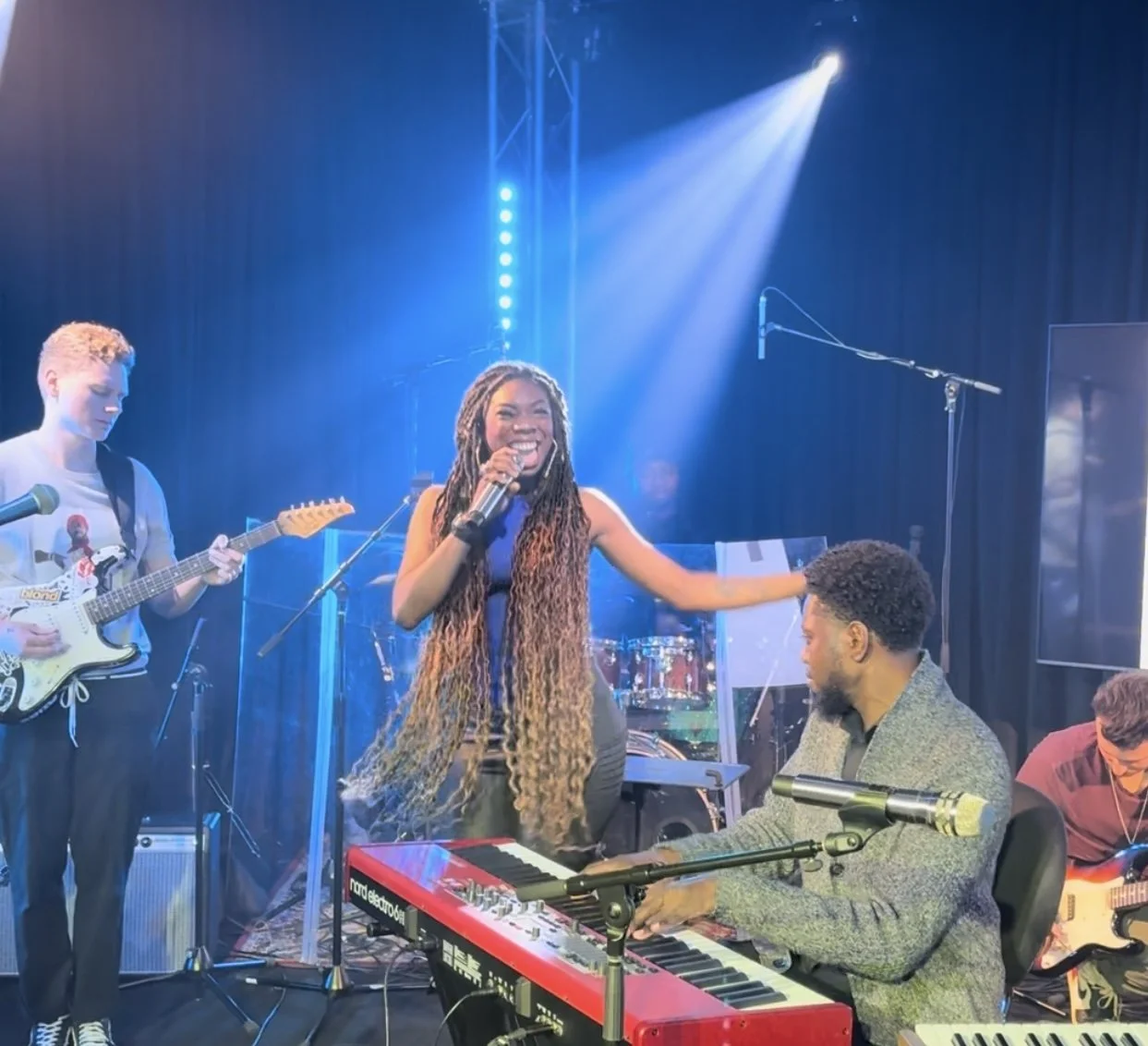 Musicians performing on stage, a woman singing into a microphone, a guitarist, and a keyboard player, under blue stage lighting.
