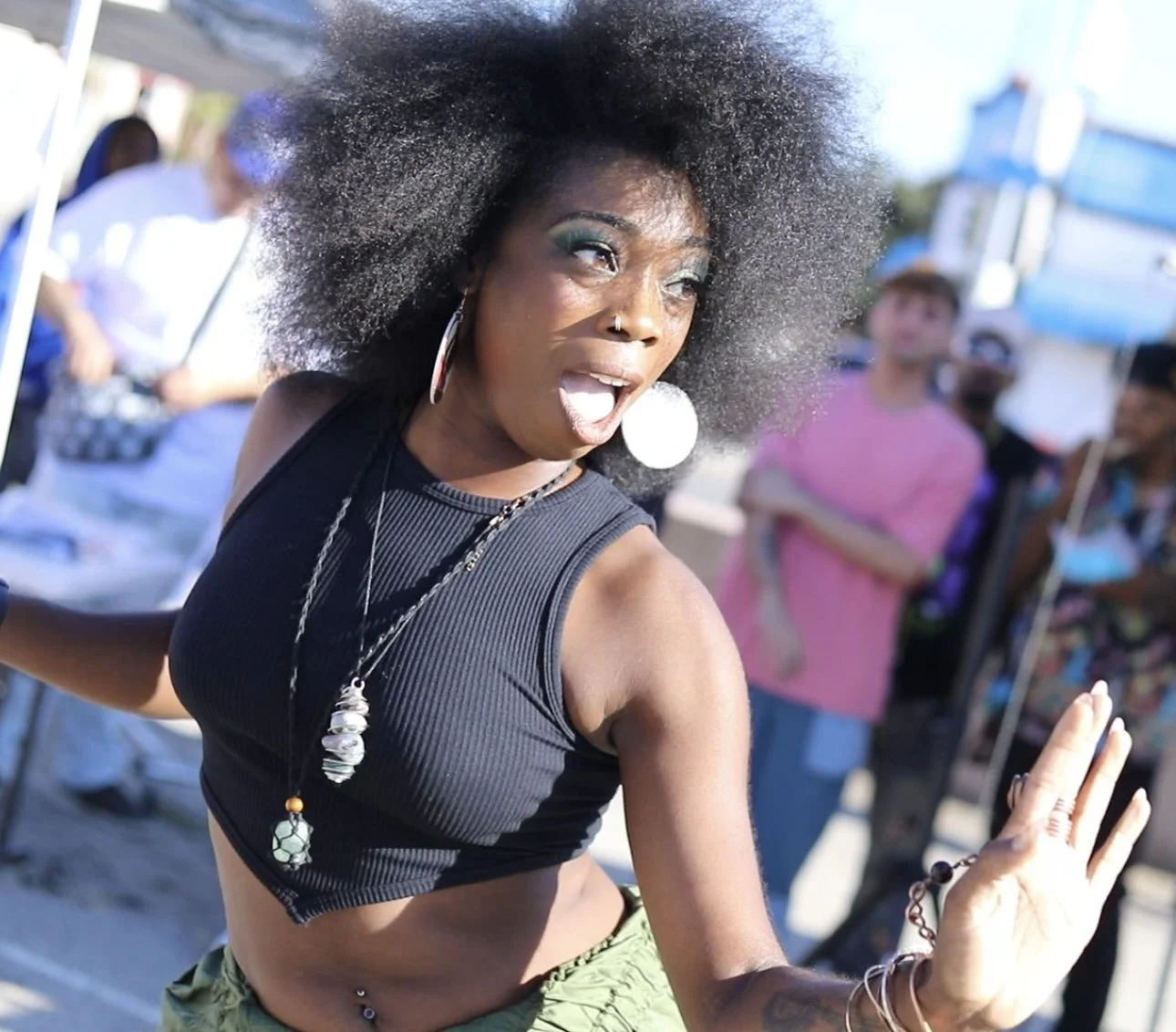 A woman dancing outdoors at a crowded event, wearing a black sleeveless crop top and green pants, with large earrings and necklaces, with her hair styled in a large afro, and surprised or excited expression on her face.
