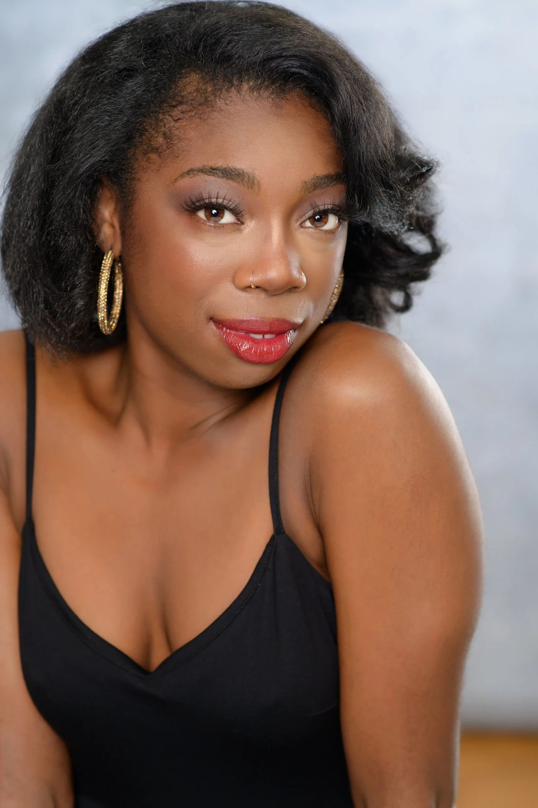 A woman with dark curly hair, hoop earrings, and red lipstick wearing a black spaghetti strap top, posing indoors with a neutral background.