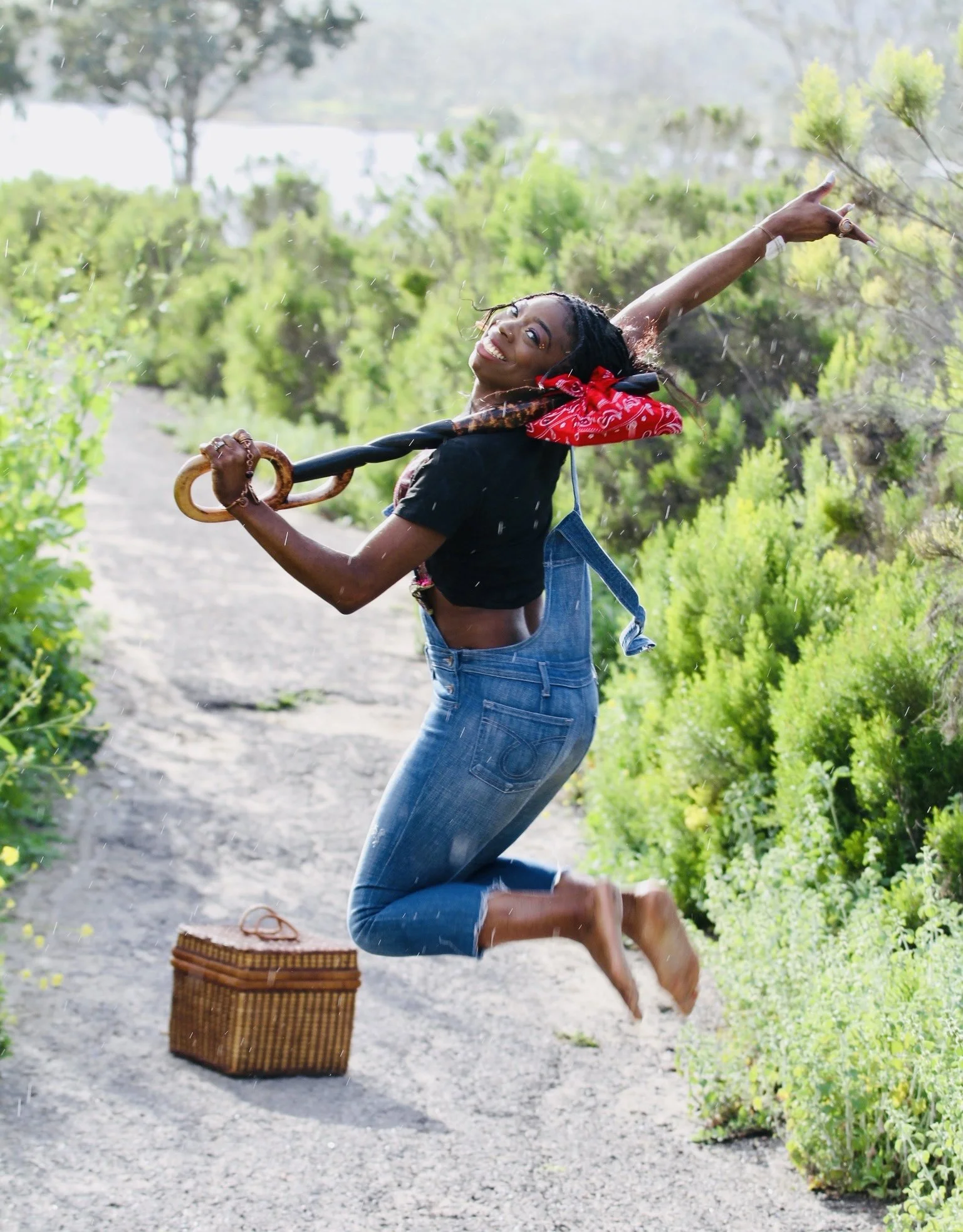 A joyful woman jumping on a dirt path surrounded by green bushes, holding a parasol, with a wicker basket nearby.