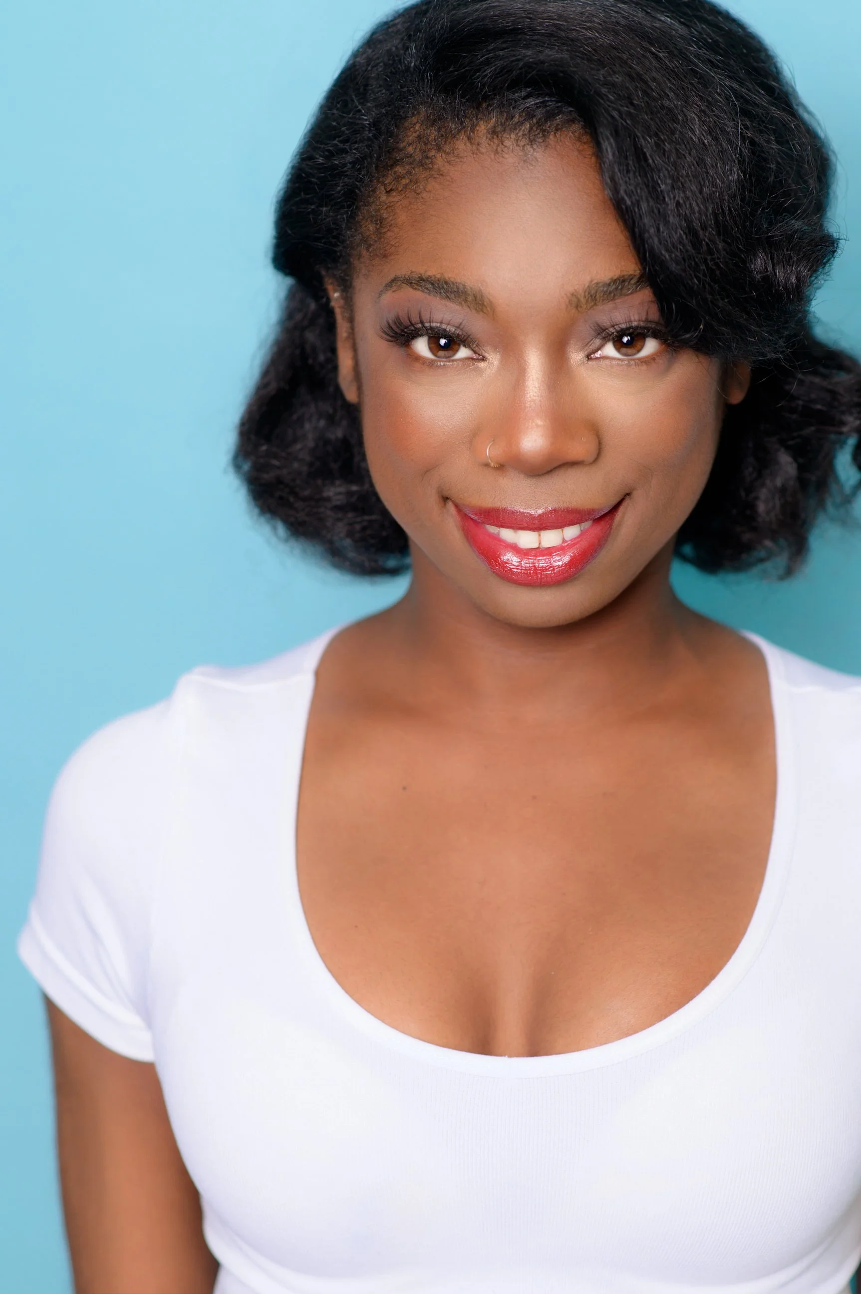 A young woman with dark skin, short curly black hair, red lipstick, and a nose ring, smiling in front of a light blue background, wearing a white shirt.