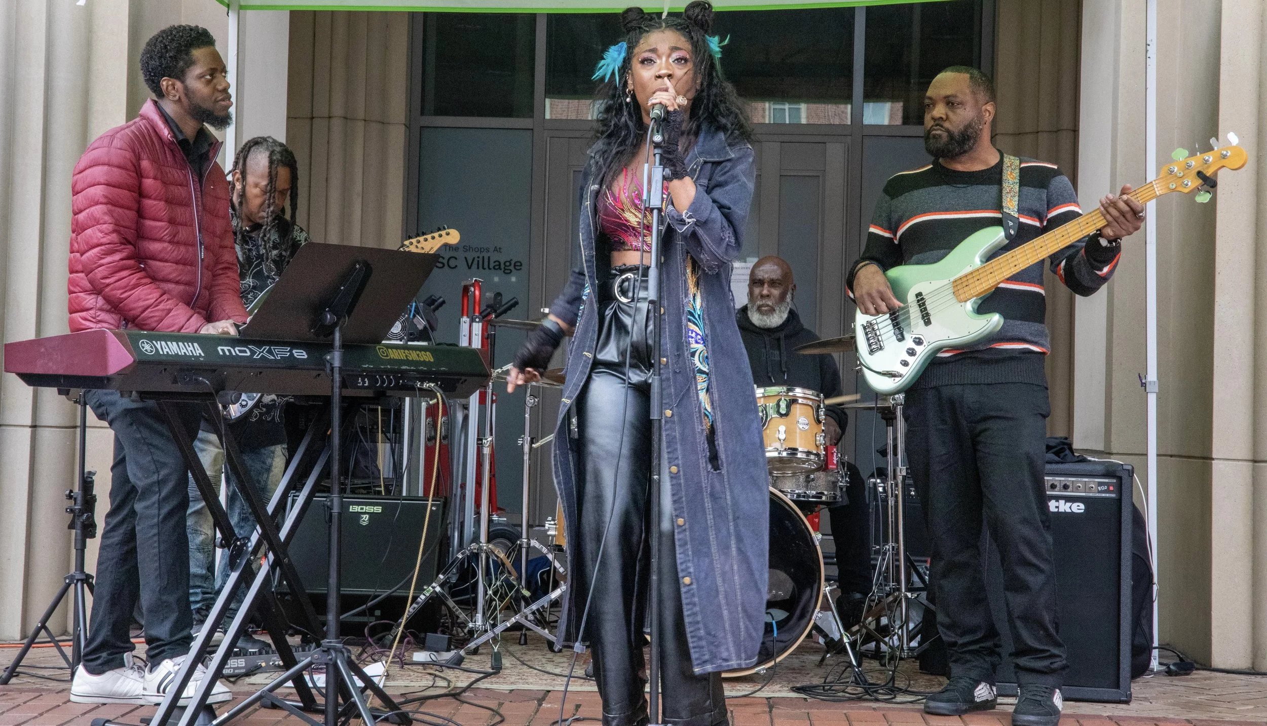 A band performing on an outdoor stage. The lead singer, a woman with textured hair and colorful feather hair accessories, is singing into a microphone. She is wearing a long denim coat and leather pants. To her left, a man with a beard is playing a w