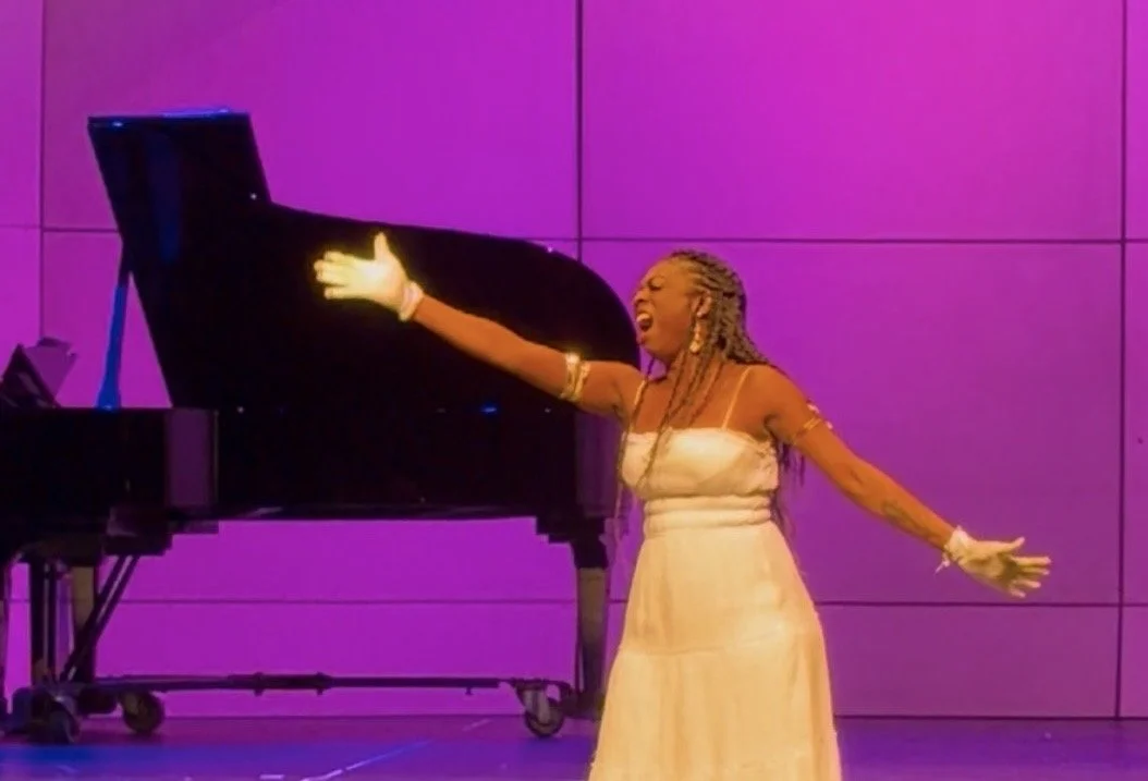 A woman with braided hair singing passionately on stage with a grand piano behind her, against a purple background.