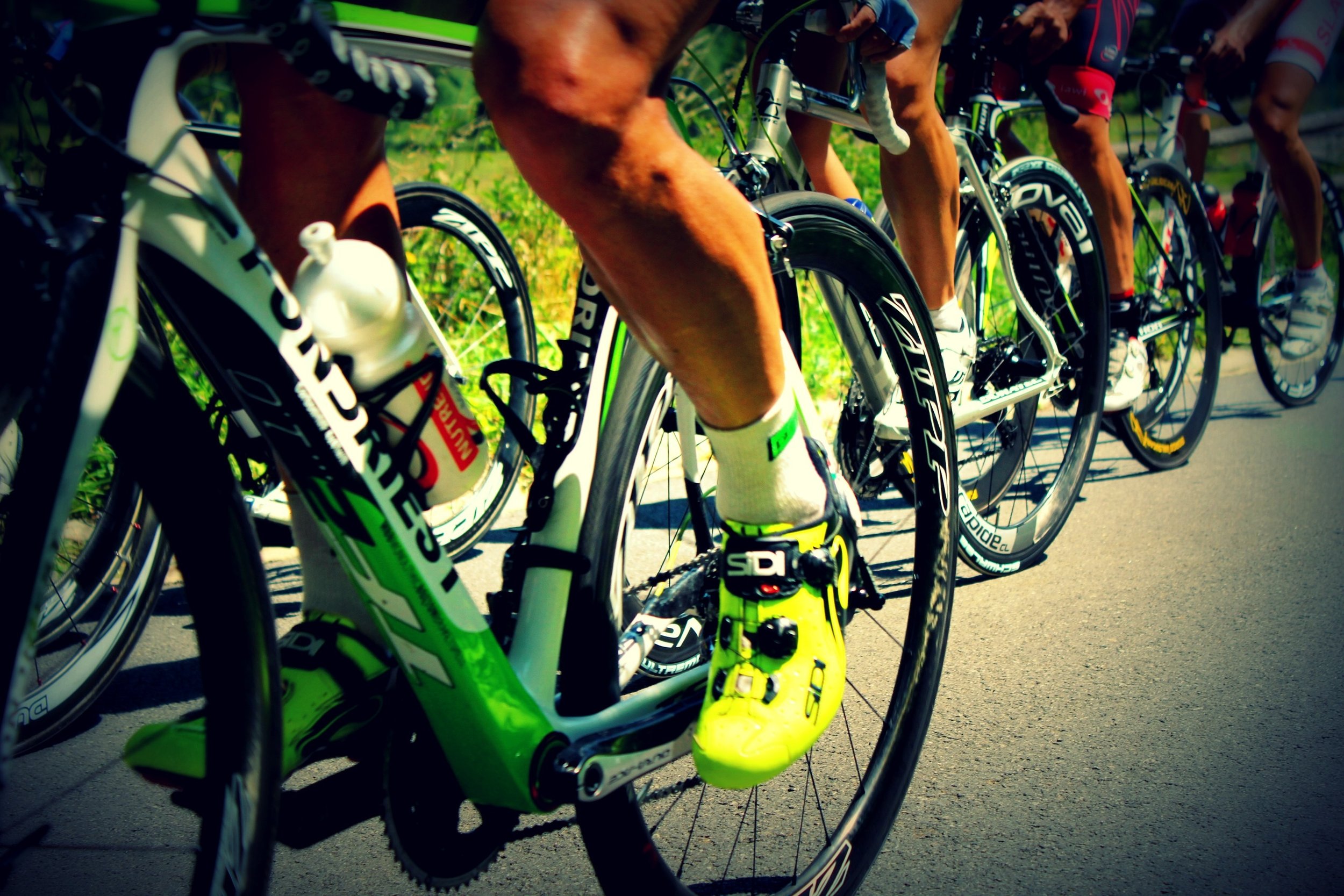 Close-up of cyclists' legs and bikes during a race, with focus on a bright yellow cycling shoe and green bike frame.