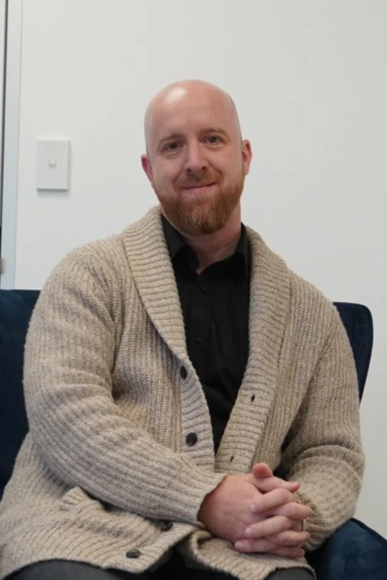 A bald man with a beard sitting on a blue chair, wearing a beige cardigan over a black shirt, with a white wall and a light switch behind him.