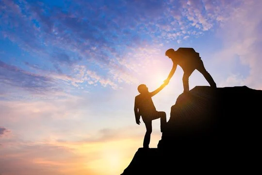 Silhouette of two people on a mountain at sunset, one helping the other climb.