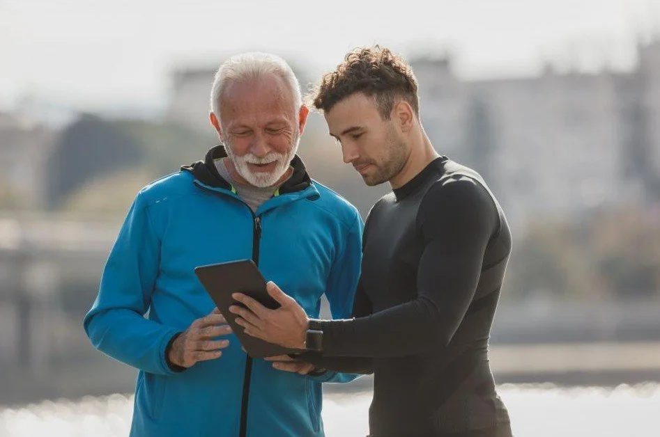 An older man and a younger man looking at a tablet together outdoors near a body of water.