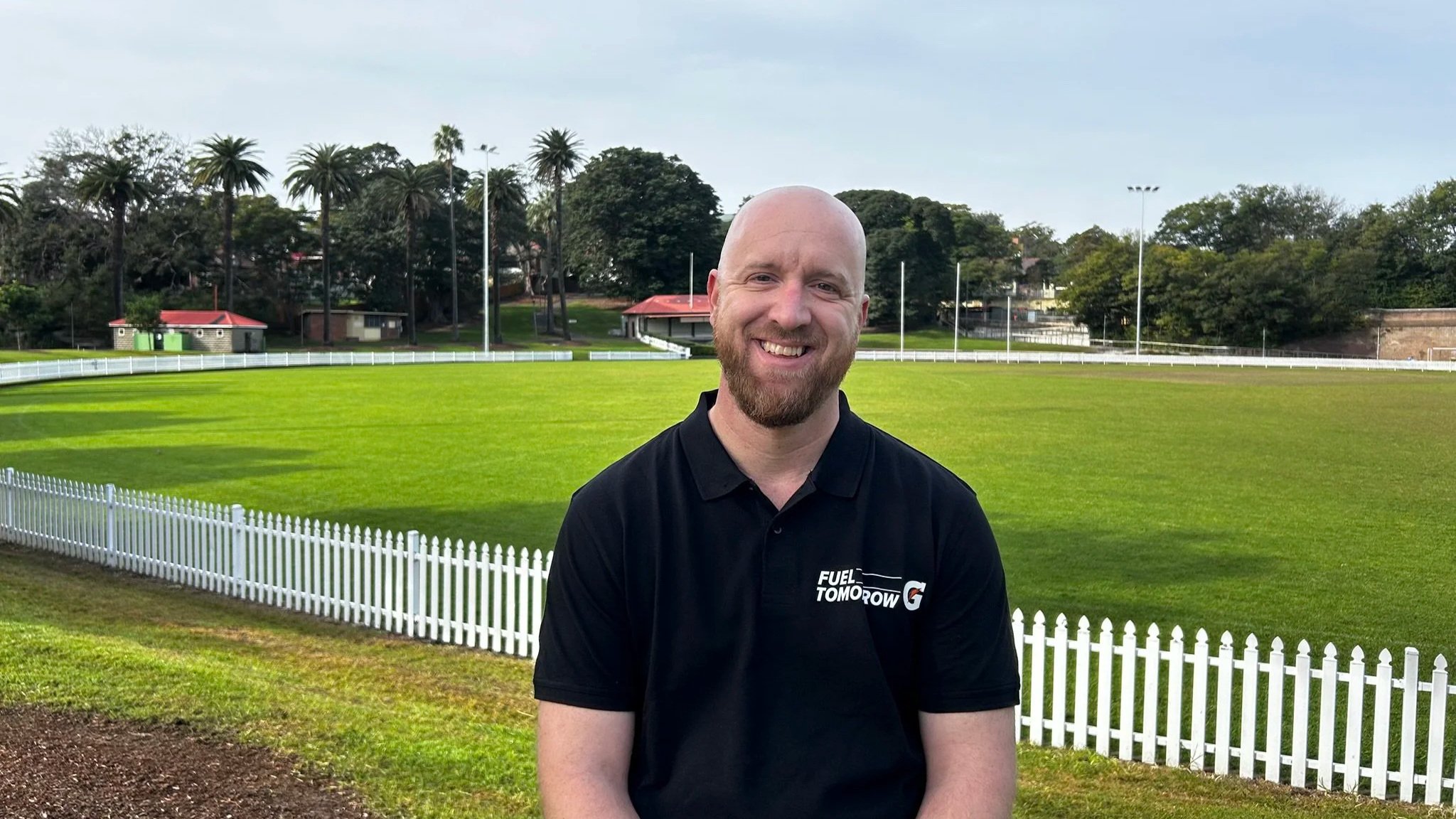 Smiling man with a bald head and beard sitting outdoors on a grassy field, with trees and a white fence in the background, wearing a black polo shirt with a logo that says Fuel Tomorrow.