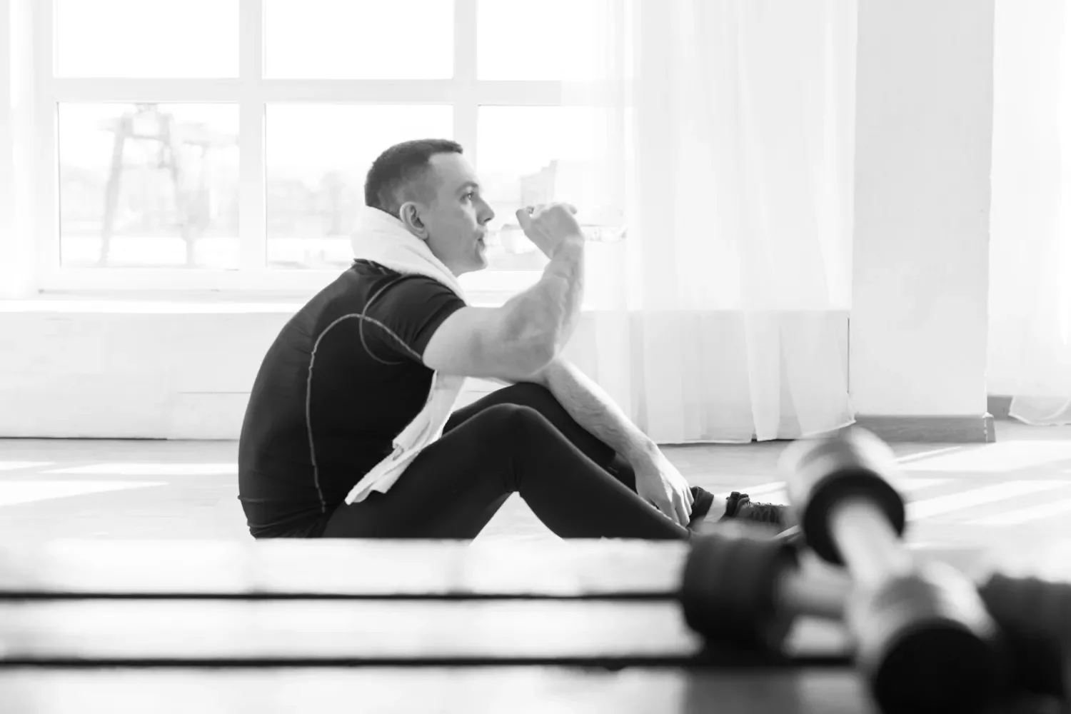 A man sitting on the floor in a gym, drinking water after workout, with dumbbells in the foreground and a large window in the background.