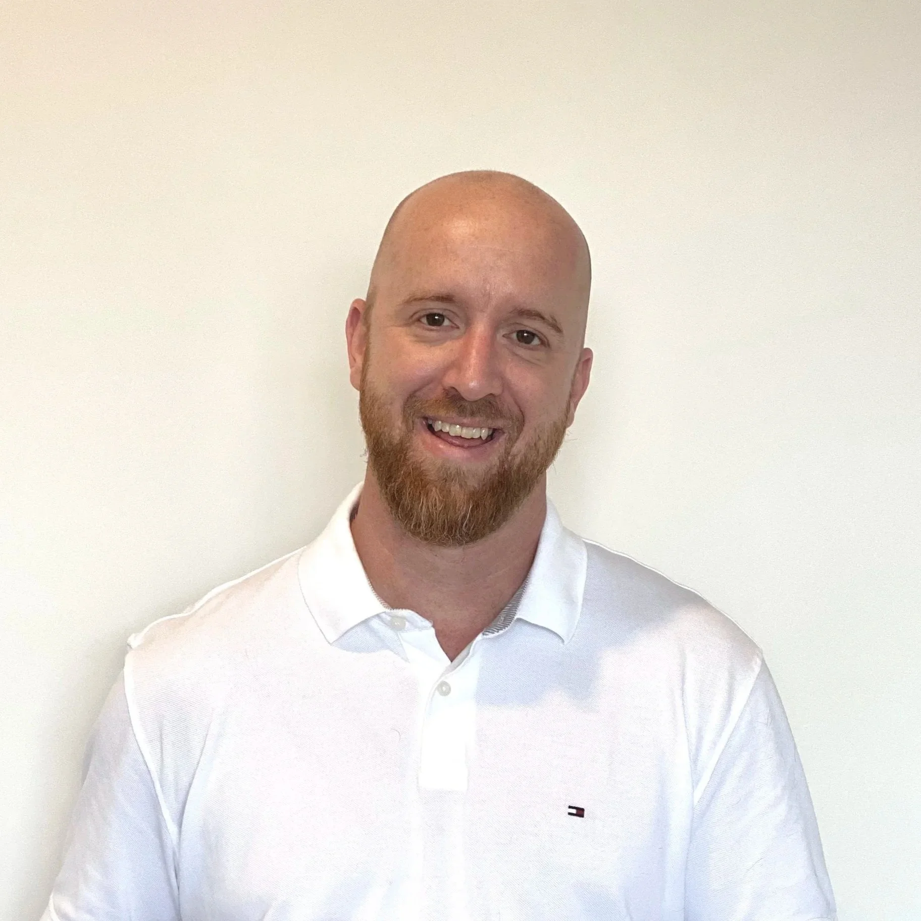 A smiling man with a beard and bald head wearing a white collared shirt standing against a plain white wall.