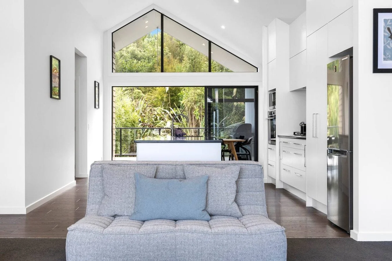 Modern open-plan living room with gray couch, kitchen island, and large windows showing greenery outside.