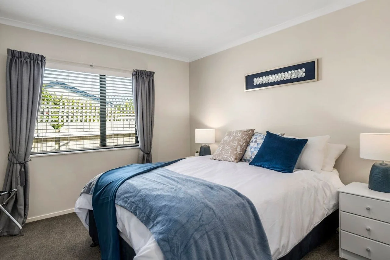 A neatly made bed with white sheets, a blue blanket, and decorative pillows in front of a window with grey curtains in a bright bedroom.