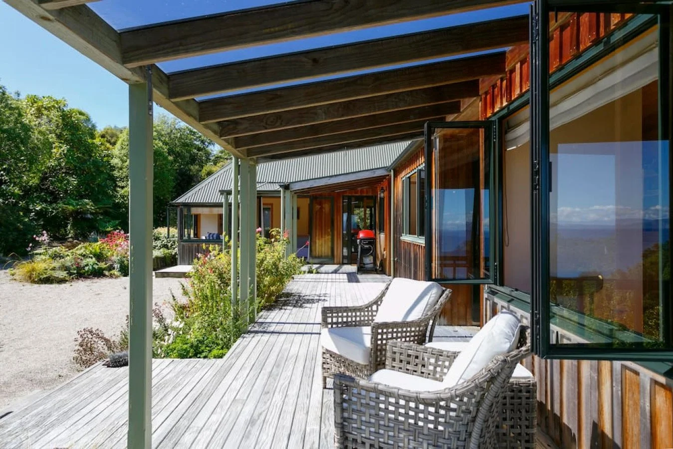 Outdoor wooden deck with wicker and cushioned chairs, partially shaded by a pergola, attached to a house with large open windows and surrounding greenery.