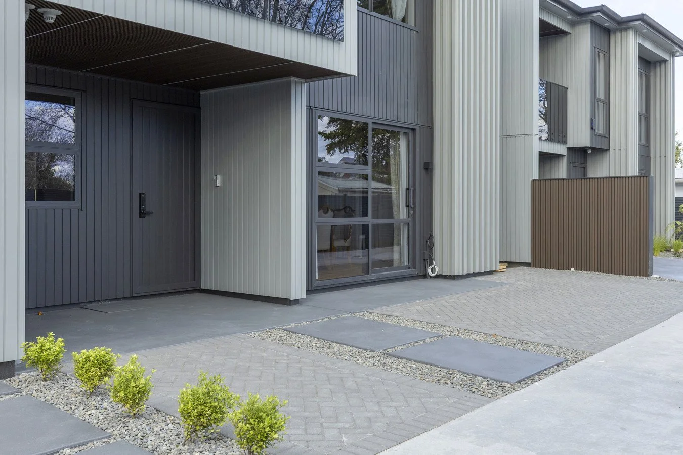 Front exterior of a modern building with horizontal and vertical metal siding in gray and beige tones, featuring a sliding glass door, small shrubs, and a paved walkway.