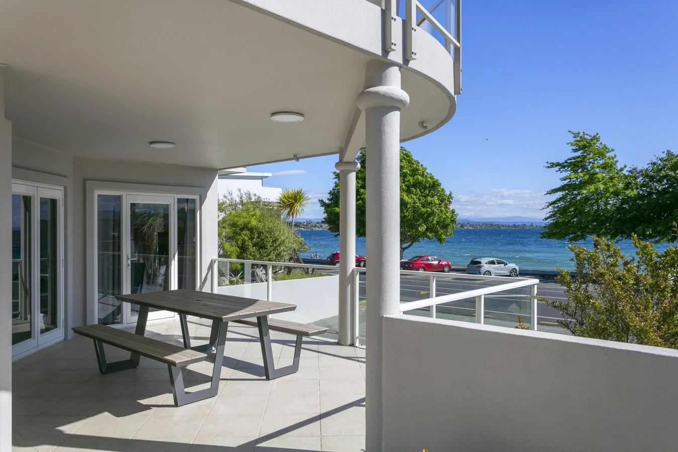 Empty balcony with a picnic table overlooking a body of water, with cars parked nearby and green trees along the shoreline, daytime with clear blue sky.