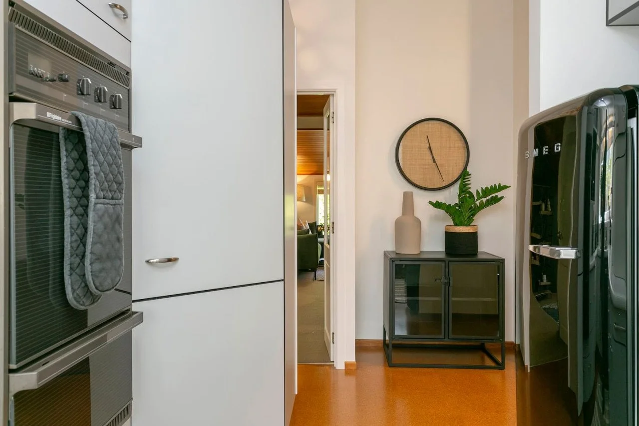 View of a modern kitchen area with a stove on the left, a white cabinet, and a hallway leading to a living room with a lamp and sofa visible. There is a black cabinet with decorative vases and a potted plant on top, a wall clock above, and a refriger