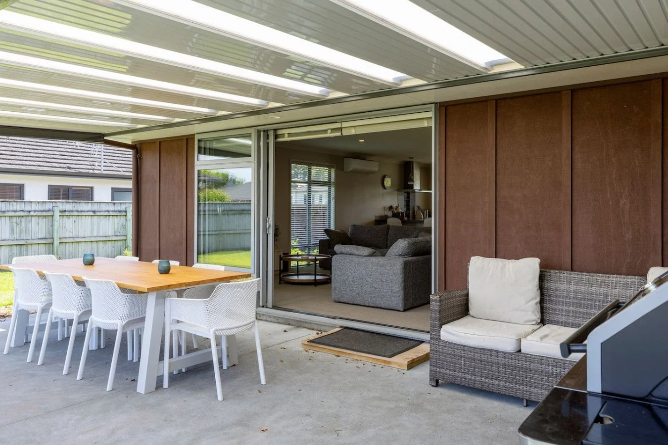 Outdoor patio area with dining table and chairs, and a wicker sofa with cushions, adjacent to a sliding glass door leading into a living room.