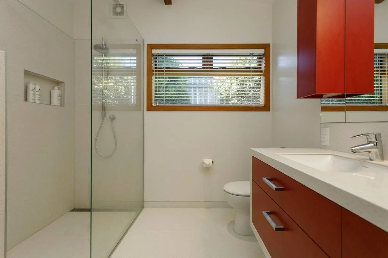 A modern bathroom with a glass-enclosed shower, a window with blinds, a white toilet, a white countertop with a sink, and red cabinetry.