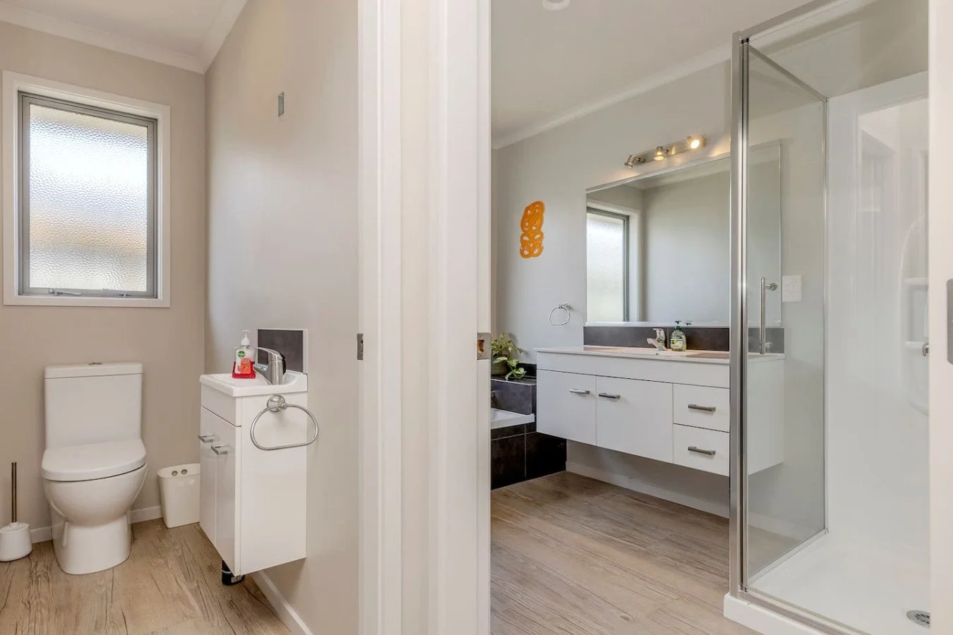 A bathroom with a toilet, small sink, and window on the left, and a vanity with a mirror and walk-in shower on the right.