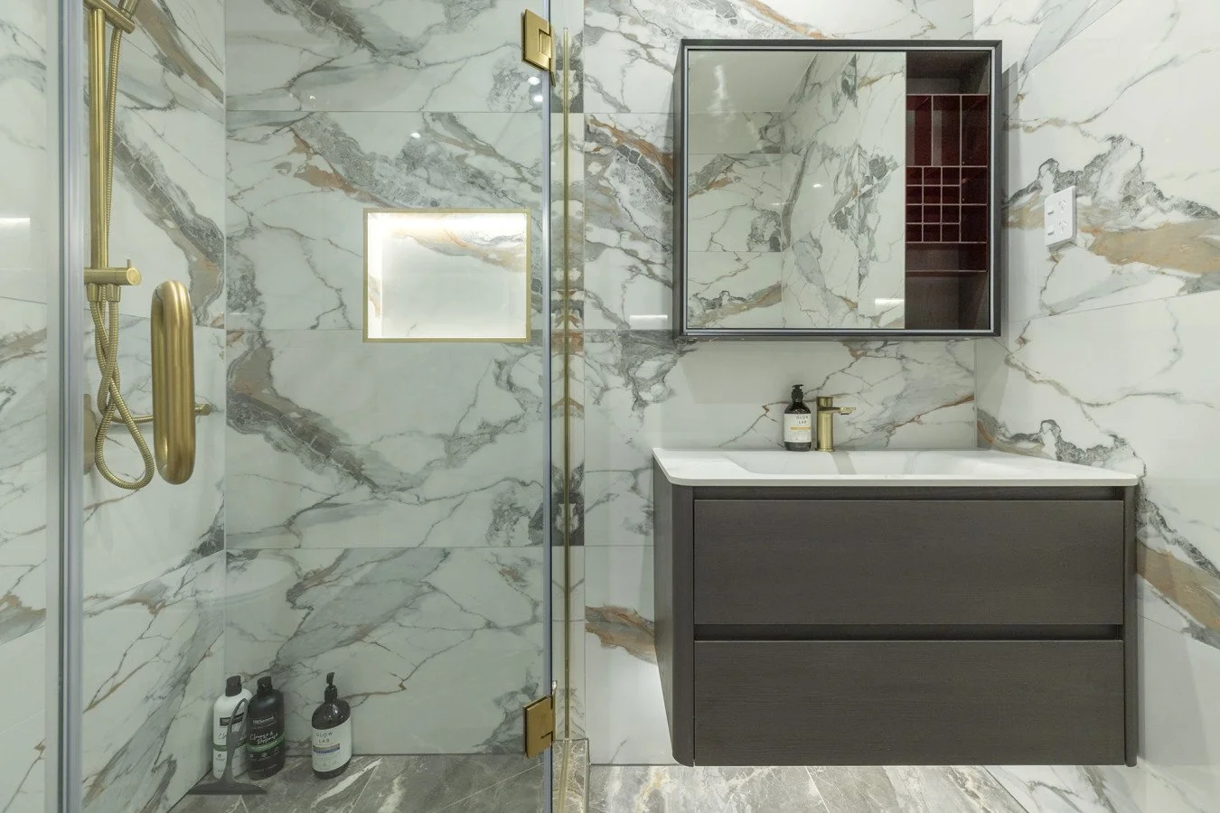 Modern bathroom with marble walls, a glass shower enclosure with gold fixtures, a dark grey vanity with a white countertop, and a mirrored cabinet.