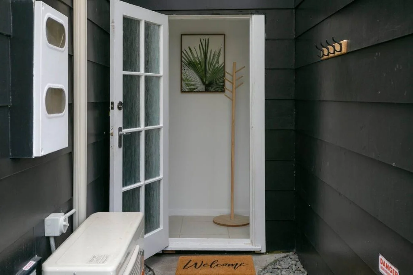Front porch entrance with black siding, a white glass-paneled door, a wood coat rack, a framed picture of a green palm leaf, a welcome mat, and various utility boxes.