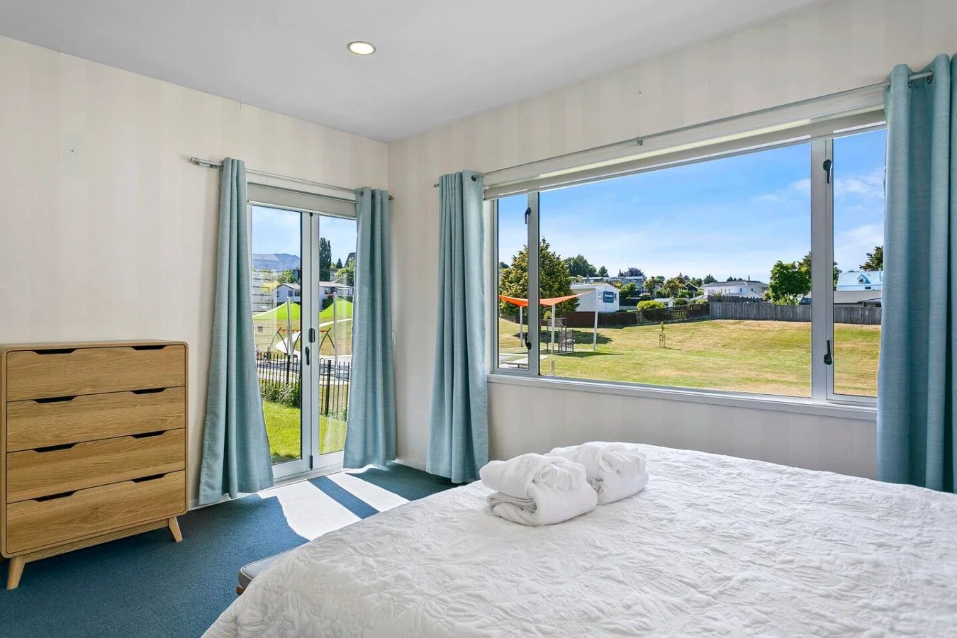 Bedroom with large windows and sliding glass door showing a backyard with a swing set and picnic table, bed with white bedding and two rolled towels, wooden dresser, and light blue curtains.