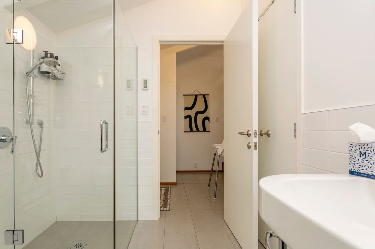 Bathroom with a glass shower enclosure, a white sink with tissue box, and an open door leading to a small hallway with a piece of abstract art on the wall.
