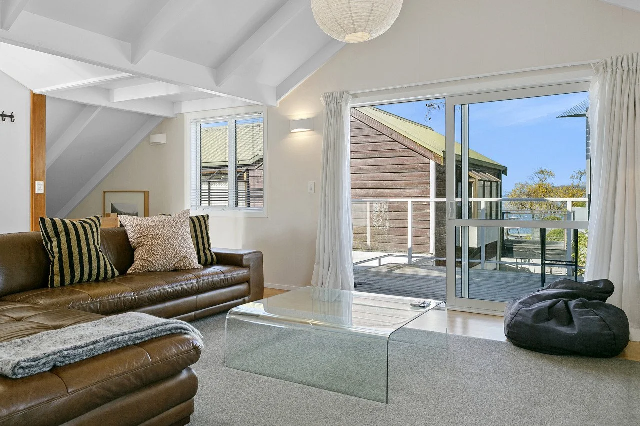 Living room with brown leather sofa, glass coffee table, white curtains, and sliding glass door leading to a balcony with outdoor view.