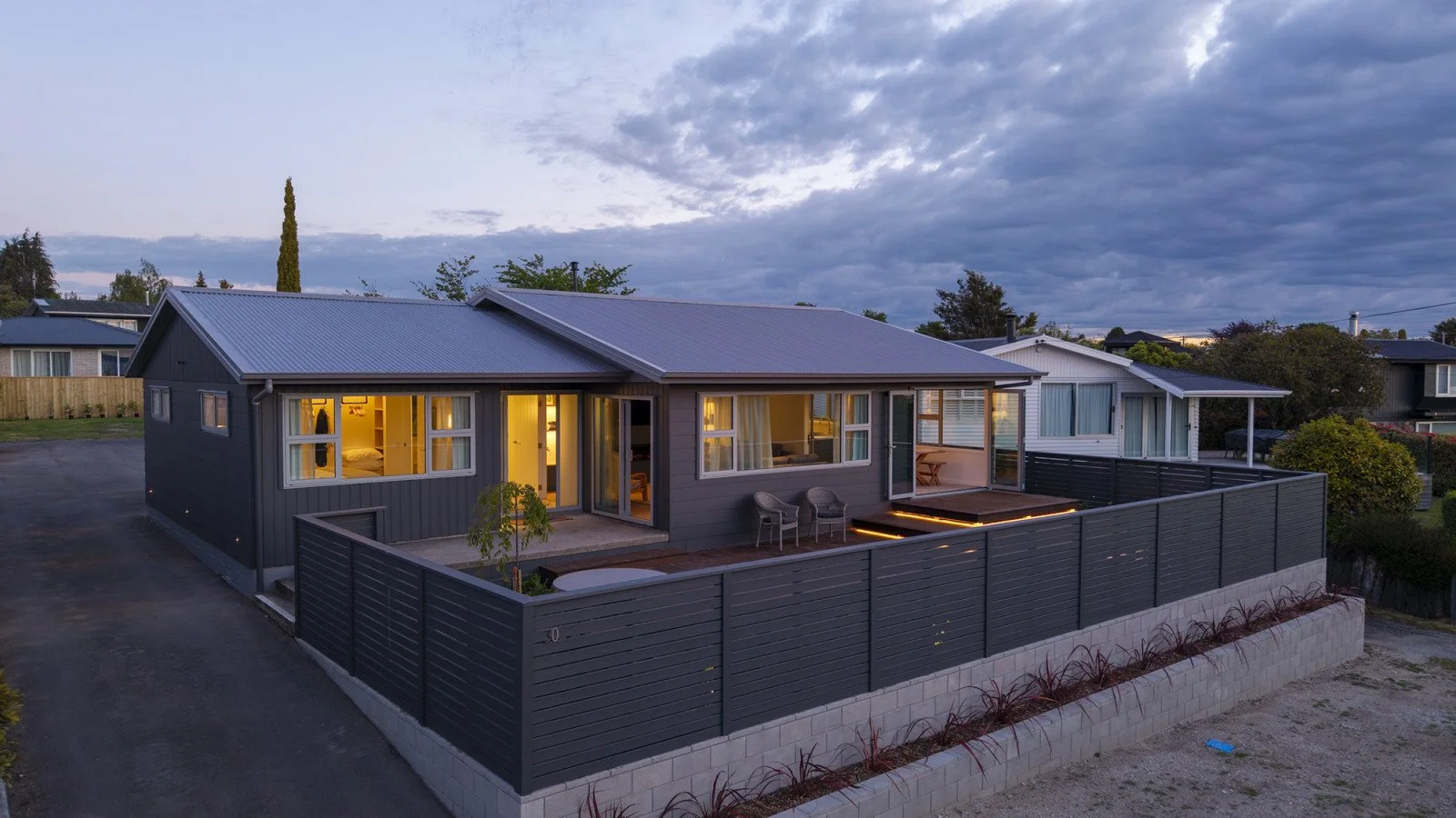 A modern house with gray siding and a metal roof, illuminated from inside, with a fenced outdoor deck, in a suburban neighborhood during dusk.
