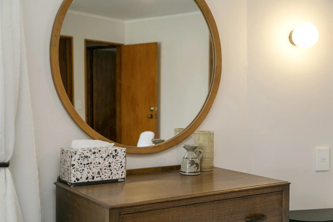 A wooden dresser with three ceramic vases and a striped tissue box, reflected in a large, round wooden-framed mirror on the wall, with a wall-mounted light fixture illuminating the scene.