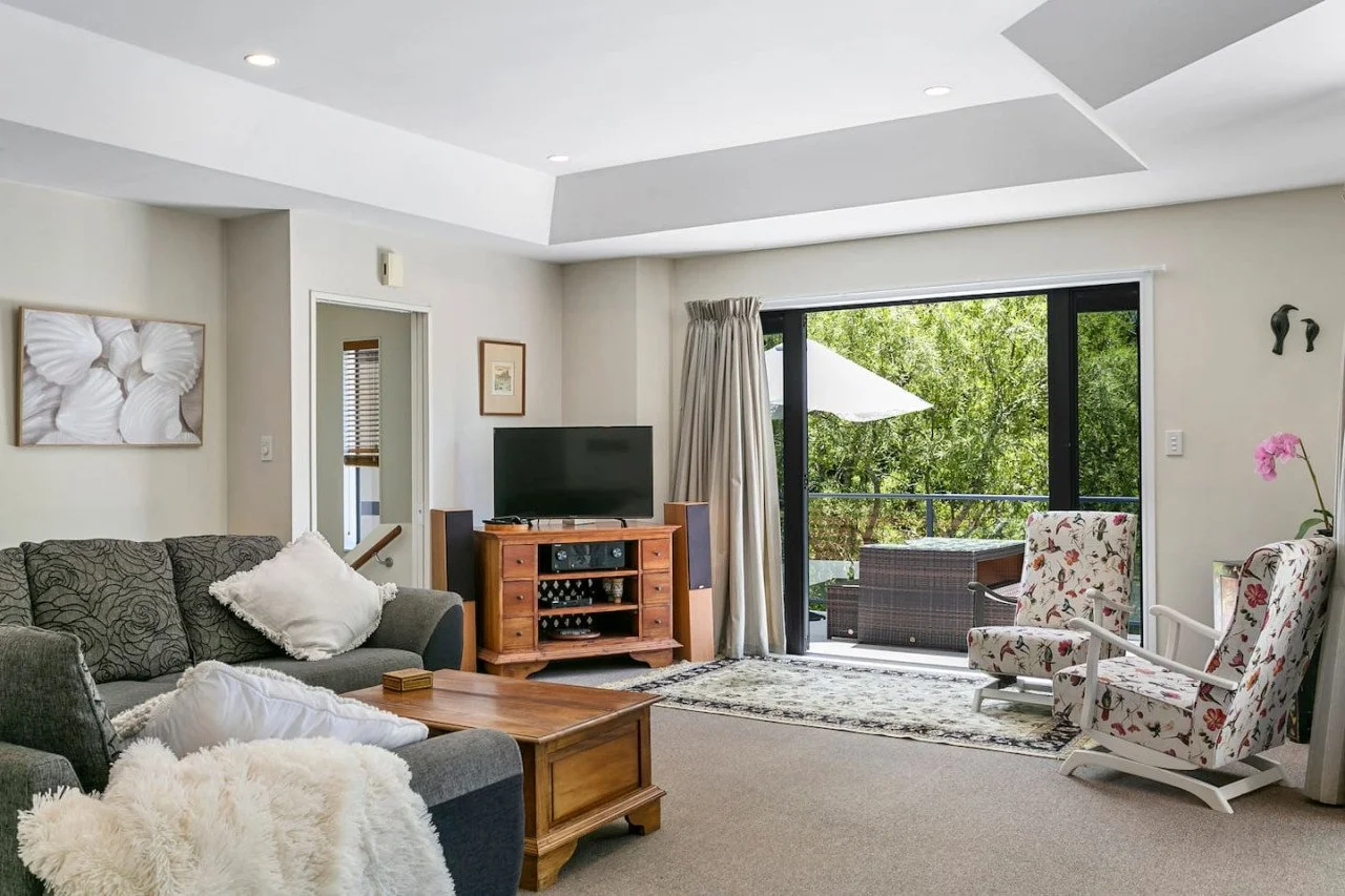 Living room with gray sofa, wooden coffee table, TV on wooden stand, floral armchairs, sliding glass door opening to balcony with outdoor furniture, and artwork on walls.