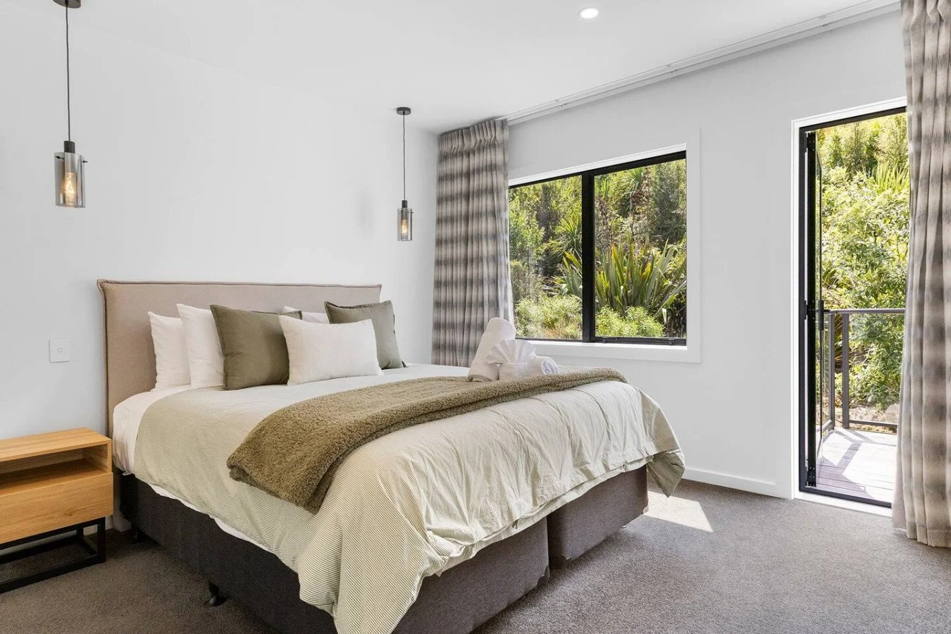 A bedroom with a large bed, beige headboard, white and green pillows, beige bedding, a wooden nightstand, hanging pendant lights, and a window with greenery outside, partially open door to a balcony.