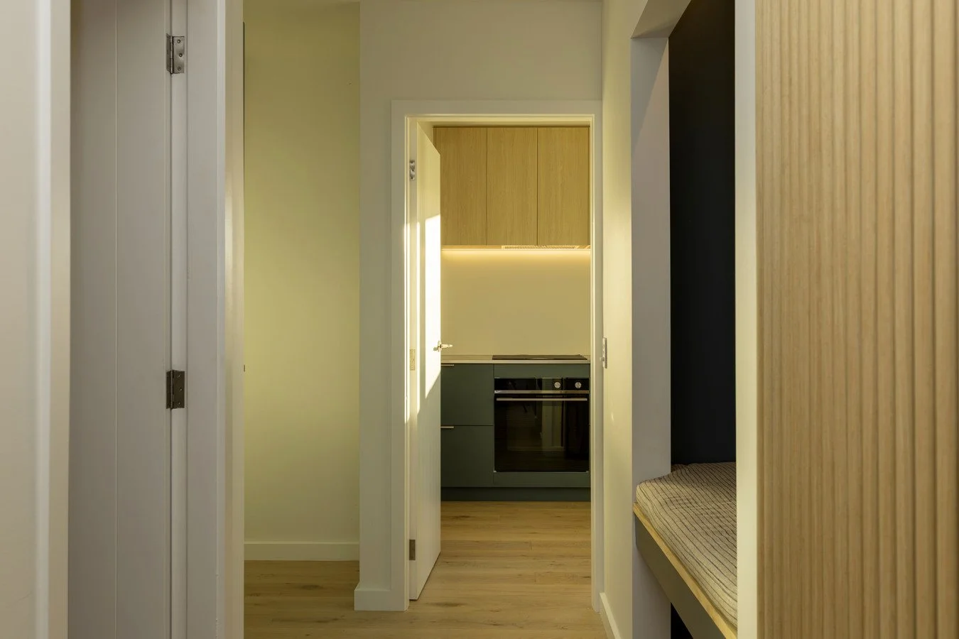 View through a doorway into a small kitchen with upper wooden cabinets, lower dark green cabinets, a built-in oven, and a beige wall with wood grain accents to the right.