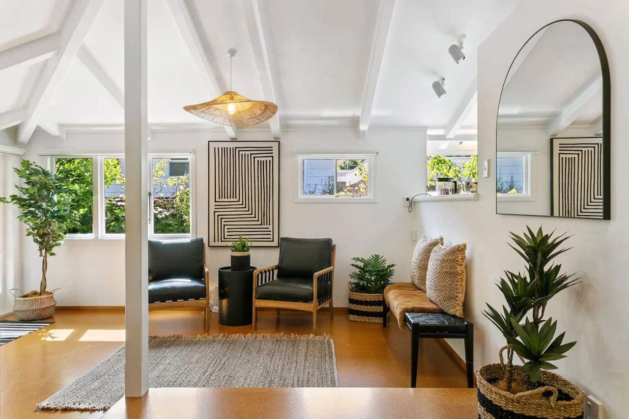 Bright living room with white walls, wood beams, and multiple windows, furnished with black armchairs, a woven bench, indoor plants, wall art, and a large mirror.