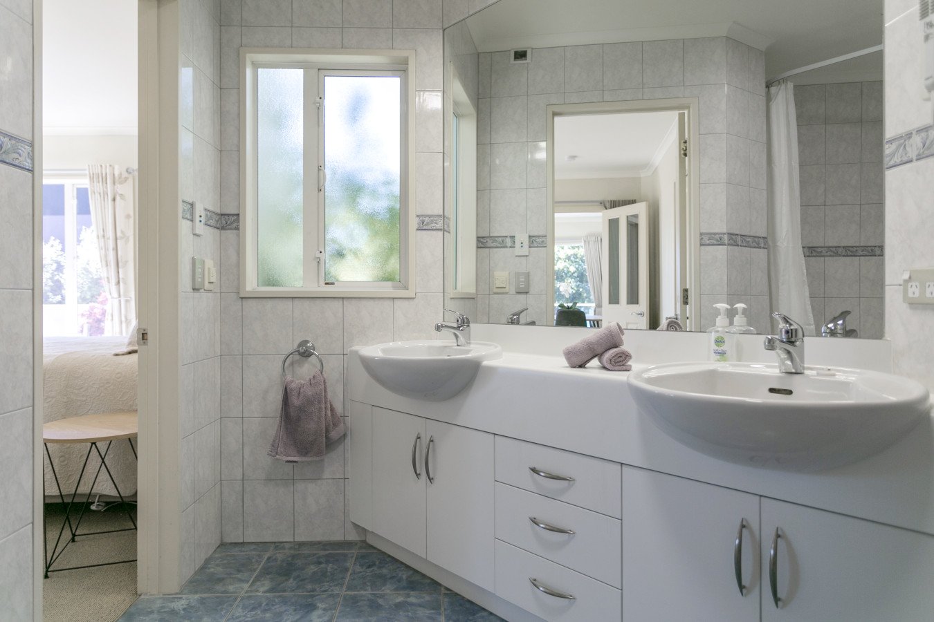 Bathroom with double sink vanity, towels, mirror, window, and door leading to a bedroom with sunlight and curtains.