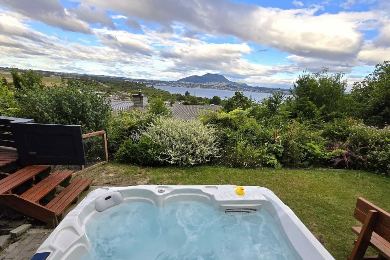 An outdoor hot tub with bubbling water on a grassy lawn, overlooking a scenic landscape with trees, a body of water, and mountains in the distance, under a partly cloudy sky.