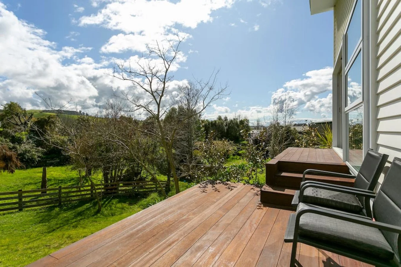A wooden deck with two chairs and a small staircase, overlooking a green backyard with trees and a partly cloudy sky.
