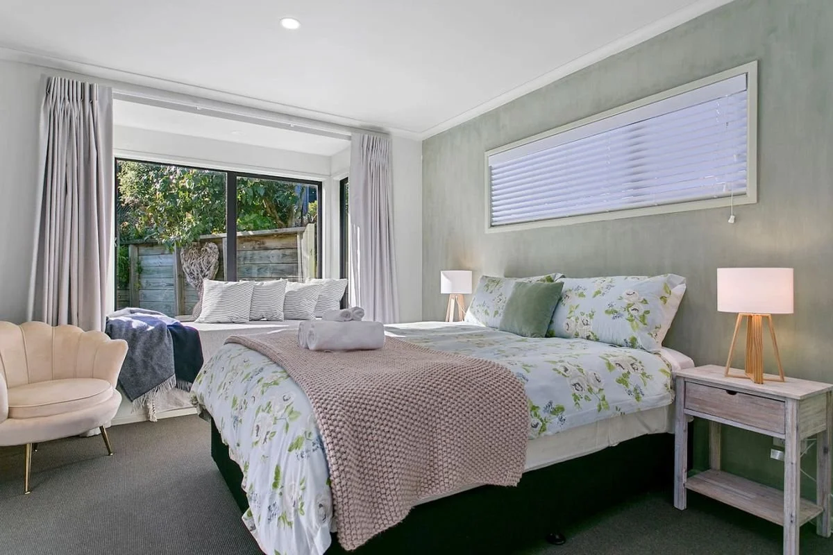 A bedroom with a large bed with floral bedding and pillows, a nightstand with a lamp, a small cream chair, and large windows with white curtains and blinds showing a garden outside.