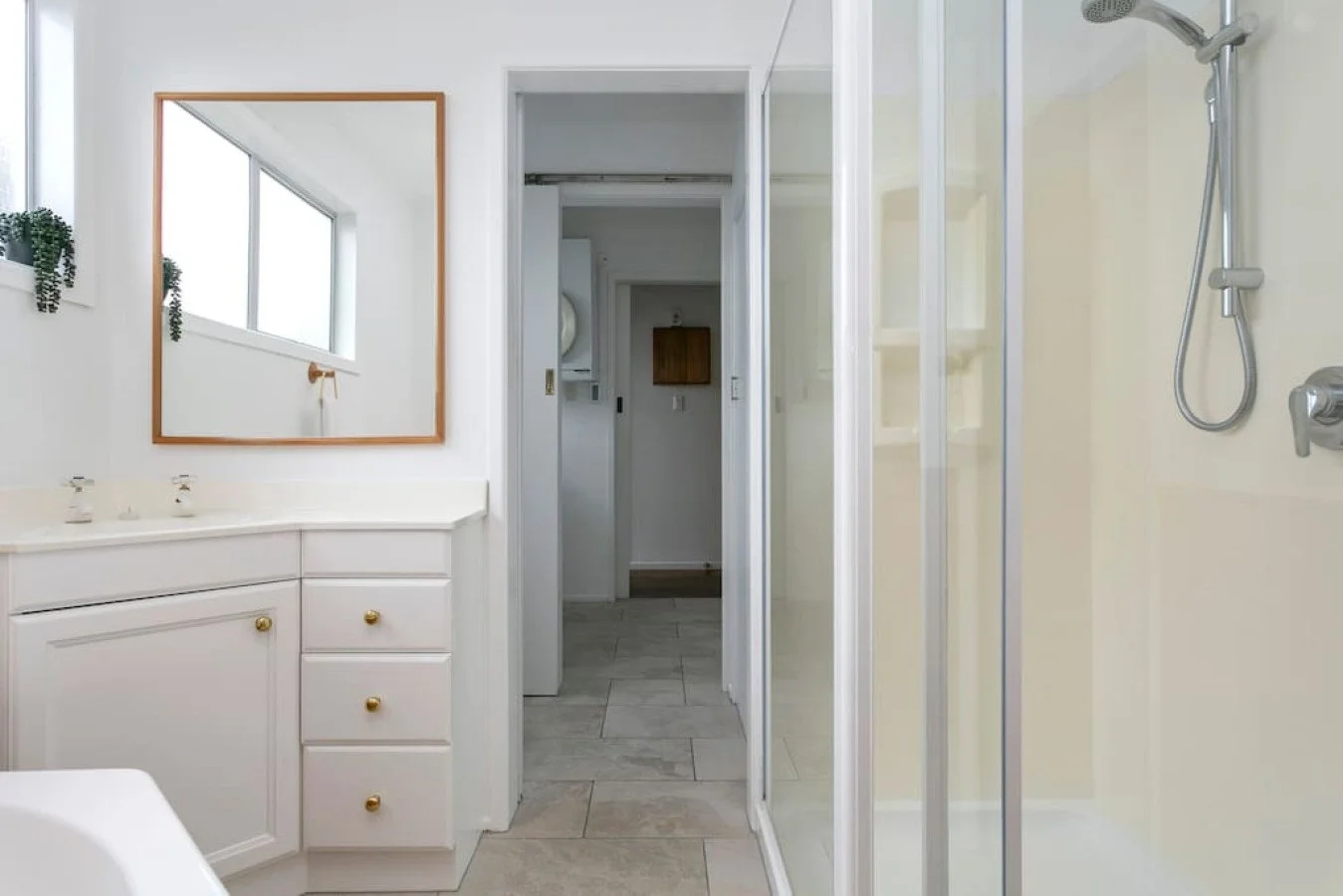 A bathroom with a white vanity countertop, a large mirror with a wooden frame, a window with hanging plants, a glass shower with a handheld showerhead, and a doorway leading to another room.