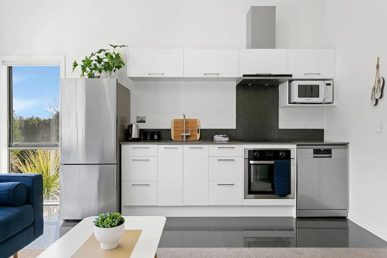 Modern kitchen with white cabinets, black countertops, stainless steel appliances, and a window showing outdoor greenery.