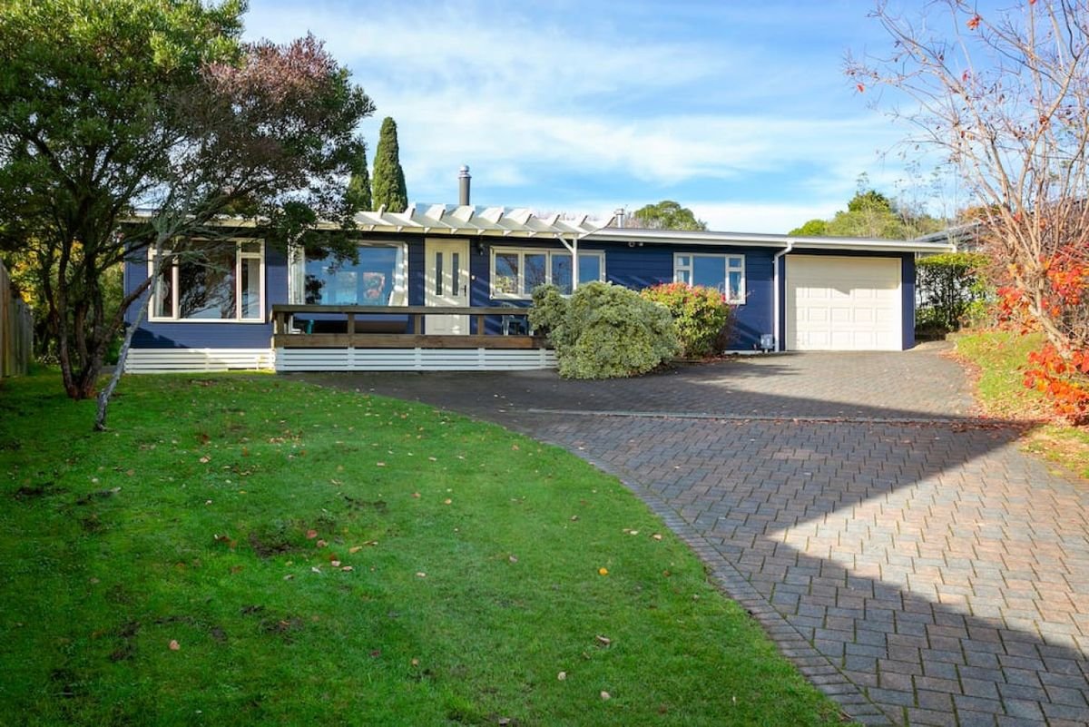 A blue single-story house with white trim, a garage, a driveway, a small porch with a railing, surrounded by trees and greenery, with a partly cloudy sky overhead.