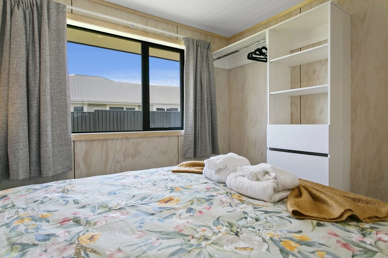 A bedroom with a large window showing a house and blue sky outside, a bed with floral bedding and towels, and a white open wardrobe.