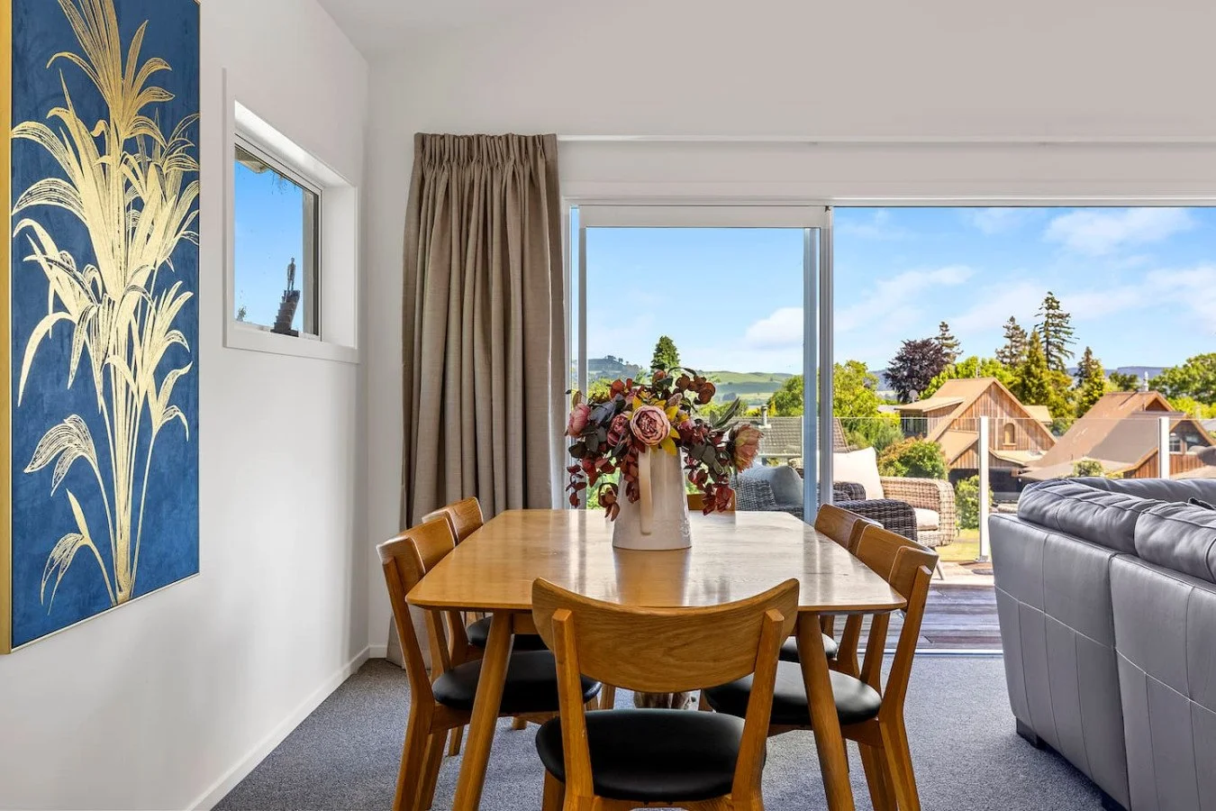 Dining area with a wooden table and six chairs, a bouquet of flowers in a pitcher in the center, sliding glass door with view of a lush outdoor landscape and neighboring houses, beige curtains, gray sofa on the right, and a blue and gold wall art on 